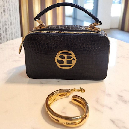 Blue handbag with gold accents on a white surface, with a gold bracelet in front.