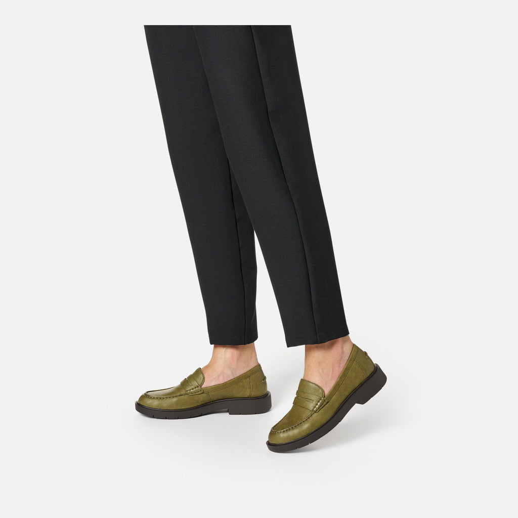 Person wearing olive green loafers and black pants on a white background