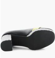 A black leather shoe shown from a side profile view, featuring a chunky black sole and a block heel with a black-and-white checkerboard pattern, accented by a small yellow detail on the side.