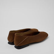 A pair of women's brown suede slip-on flats with black soles, shown from a three-quarters rear angle on a light gray background.
