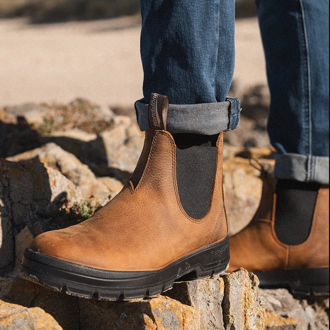 A person wearing brown leather Chelsea-style boots with black elastic side panels, shown outdoors on rocks with blue jeans cuffed above the ankles.